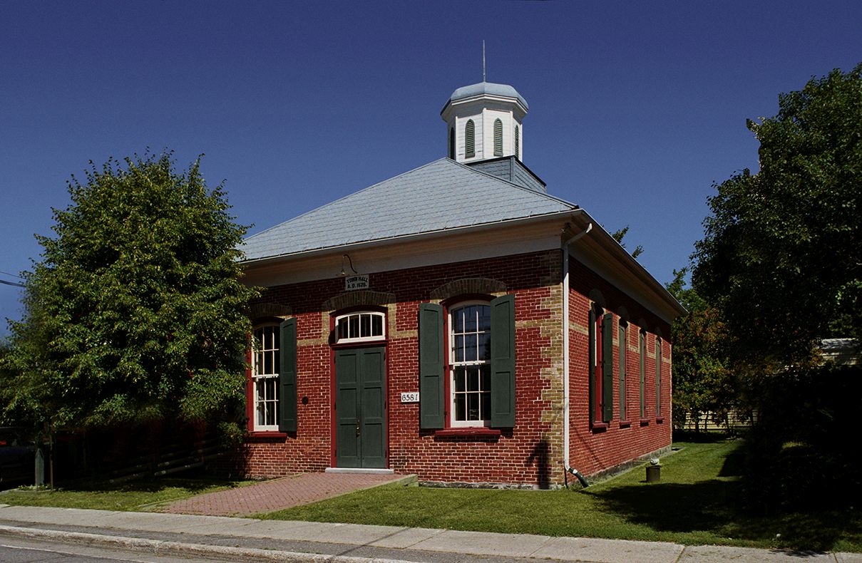 Exterior view of a one-storey brick-faced modest town hall with careful detailing and a cupola, built in 1876.